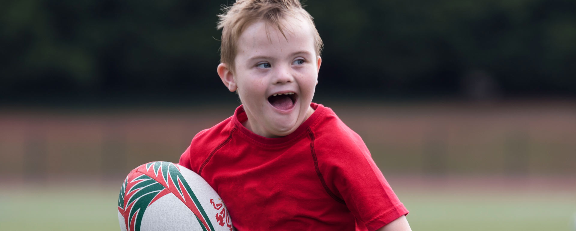 A young boy with Down's syndrome is playing running rugby and is smiling broadly.